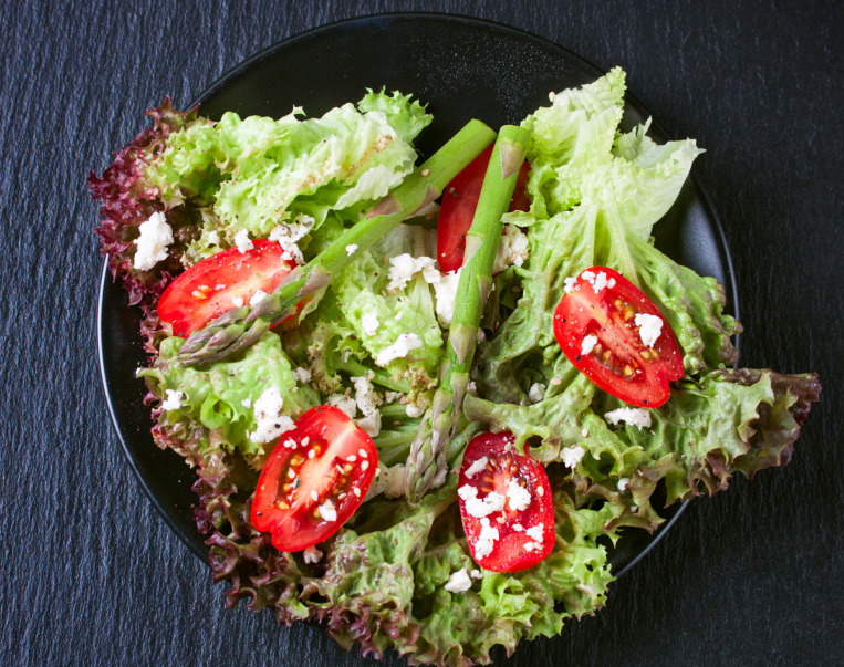 Homemade fresh salad with asparagus, tomatoes, cheese, salad leaves and cheese on a dark background, top view